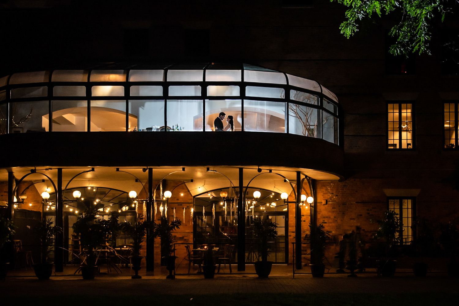 A stunning wedding venue glows warmly at night with silhouetted couple visible through large windows.
