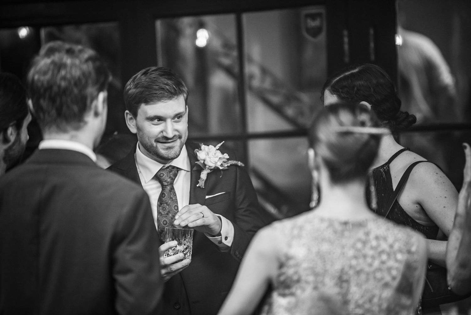Groom in navy suit with boutonniere laughing with wedding guests in black and white photo.