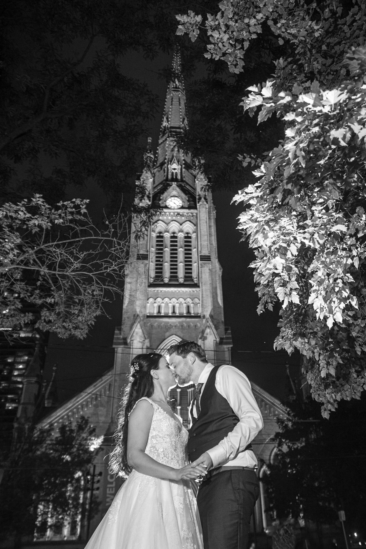 Black and white photo of bride and groom embracing before a lit Gothic church tower at night.