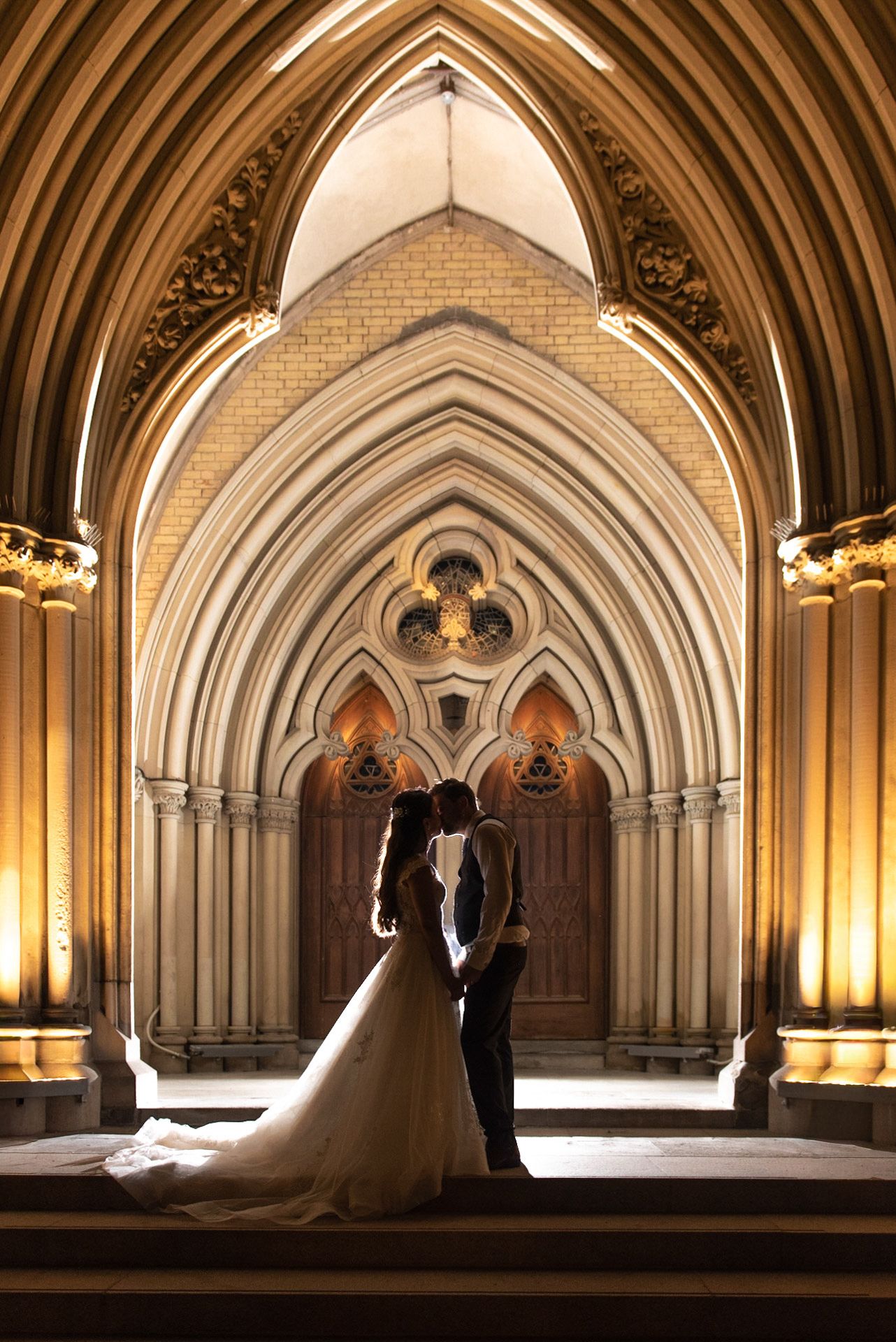 Couple shares a kiss beneath ornate Gothic cathedral arches glowing with warm amber uplighting.