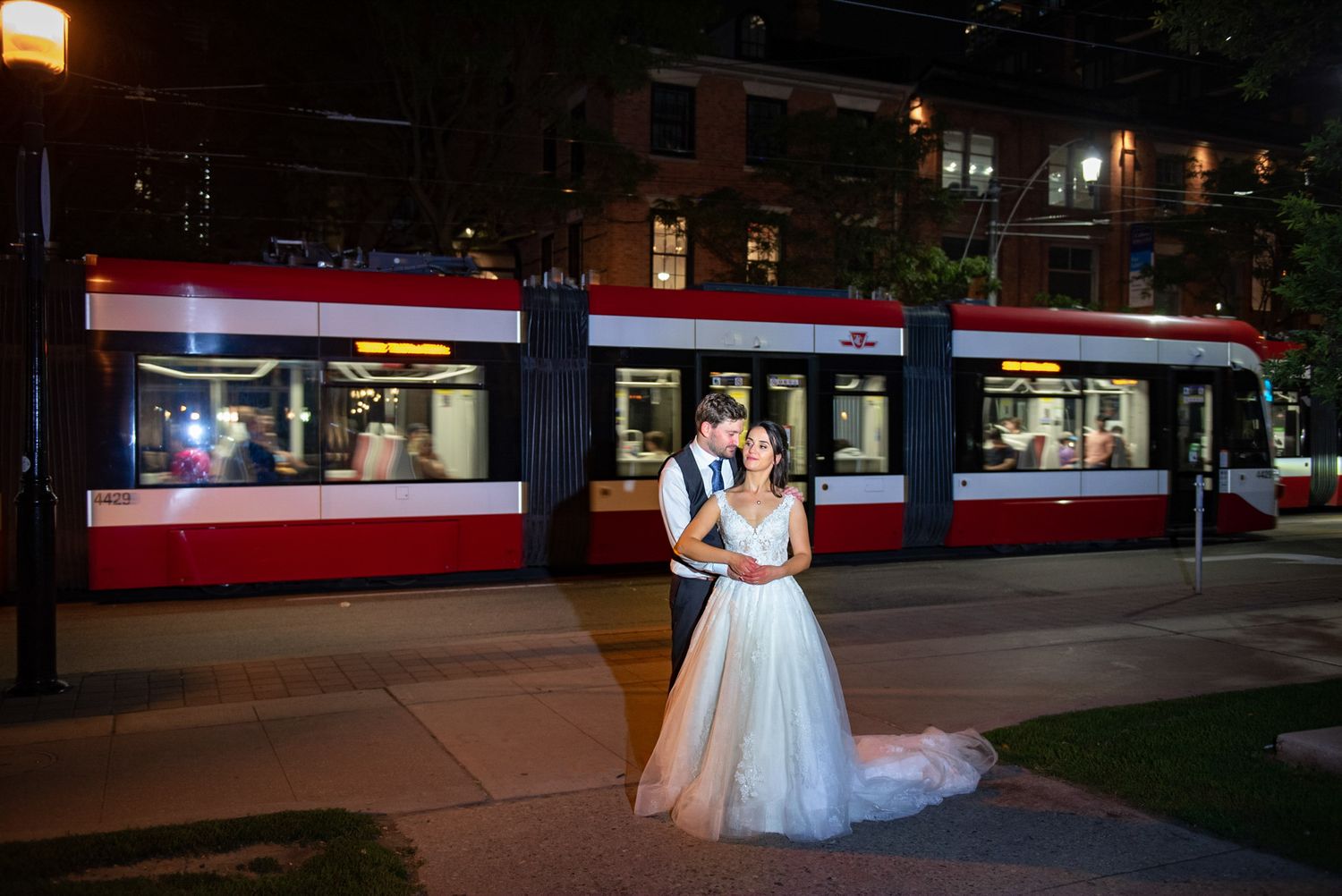 Newlyweds posing romantically in front of a red streetcar on a city street at night.