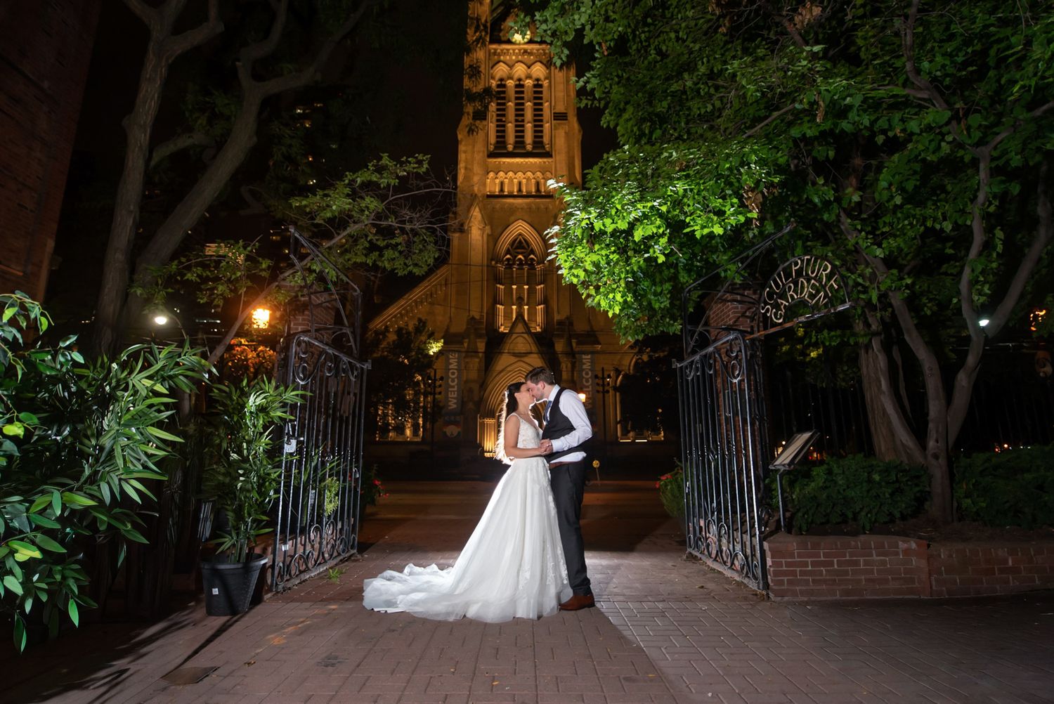 Bride and groom embrace romantically at night with a beautifully lit Gothic stone church behind them.