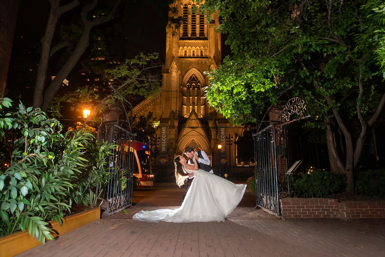 Groom dips bride dramatically outside illuminated Gothic church gates during nighttime wedding photos.