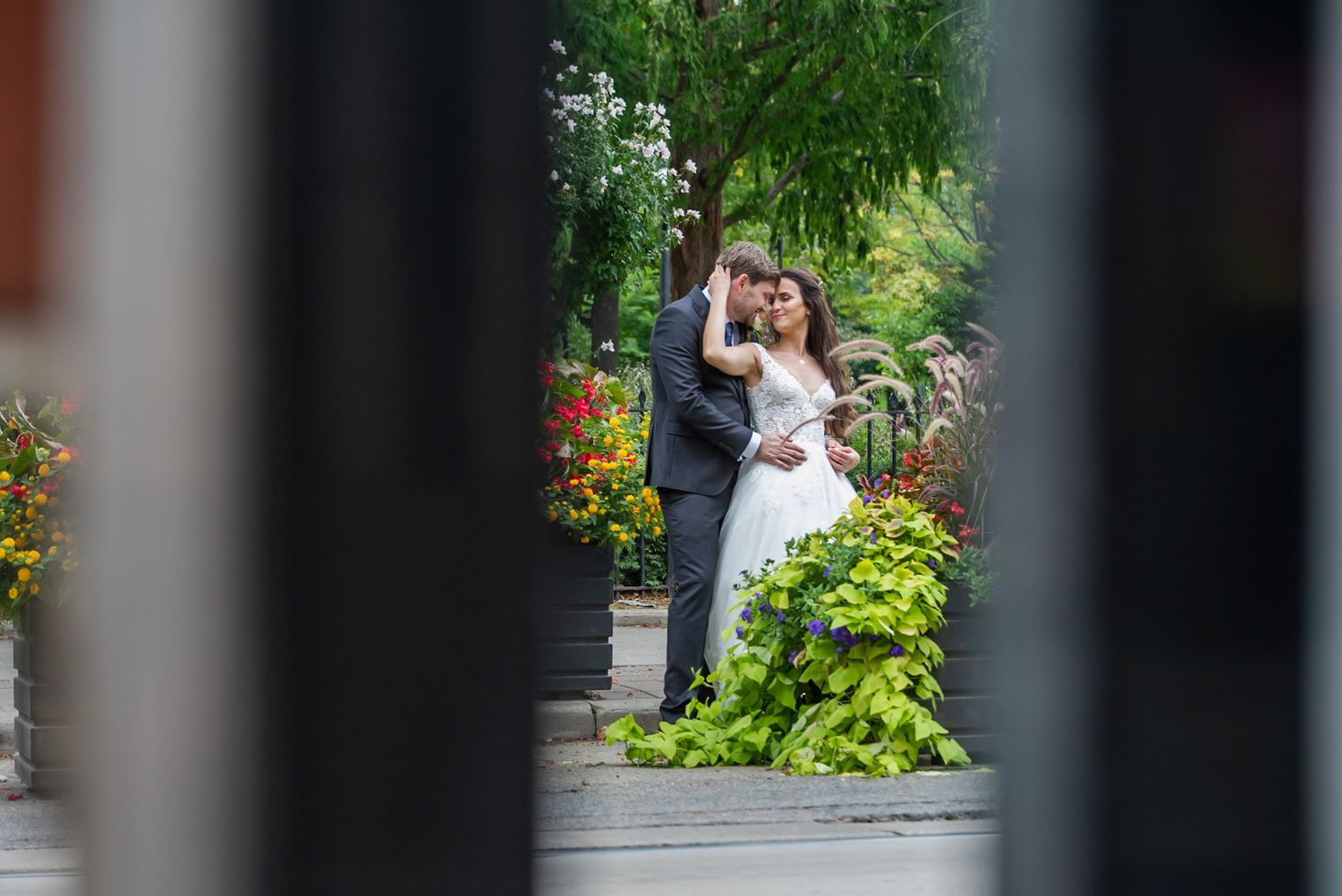 Bride and groom embracing in a colorful garden, photographed through decorative iron fence posts.