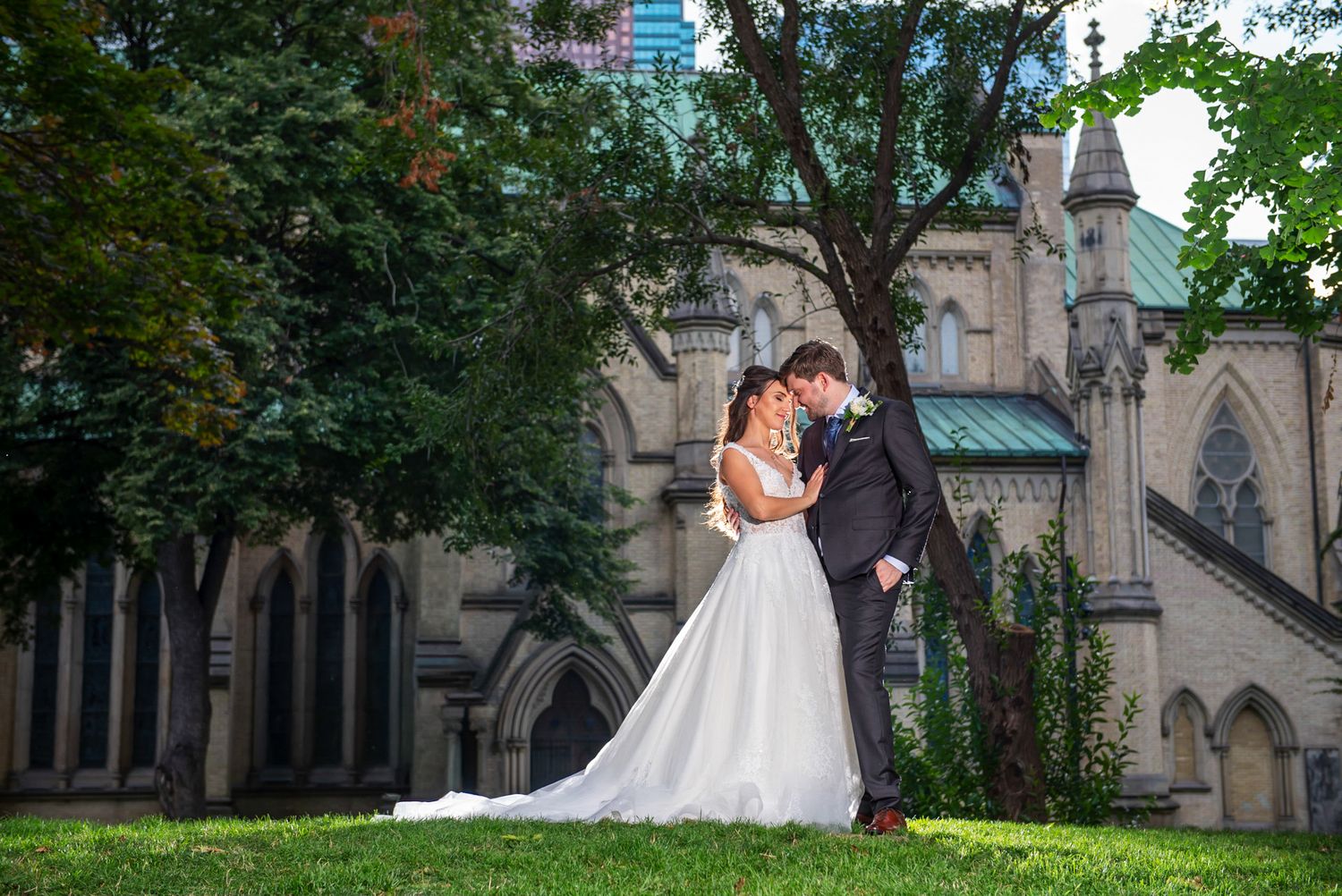 Bride and groom share an intimate moment on the grass in front of a stunning Gothic stone church.