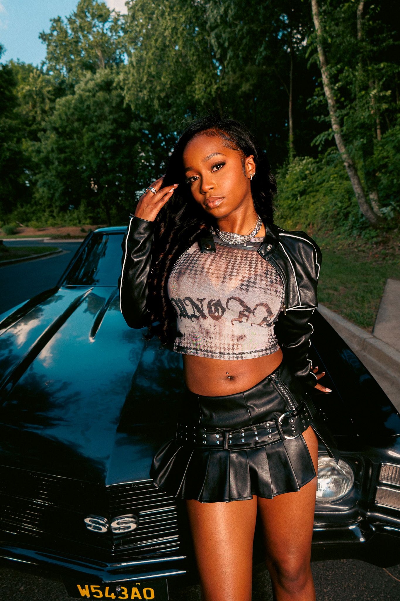Young woman in leather jacket, crop top, and mini skirt poses confidently in front of a classic black SS muscle car outdoors.