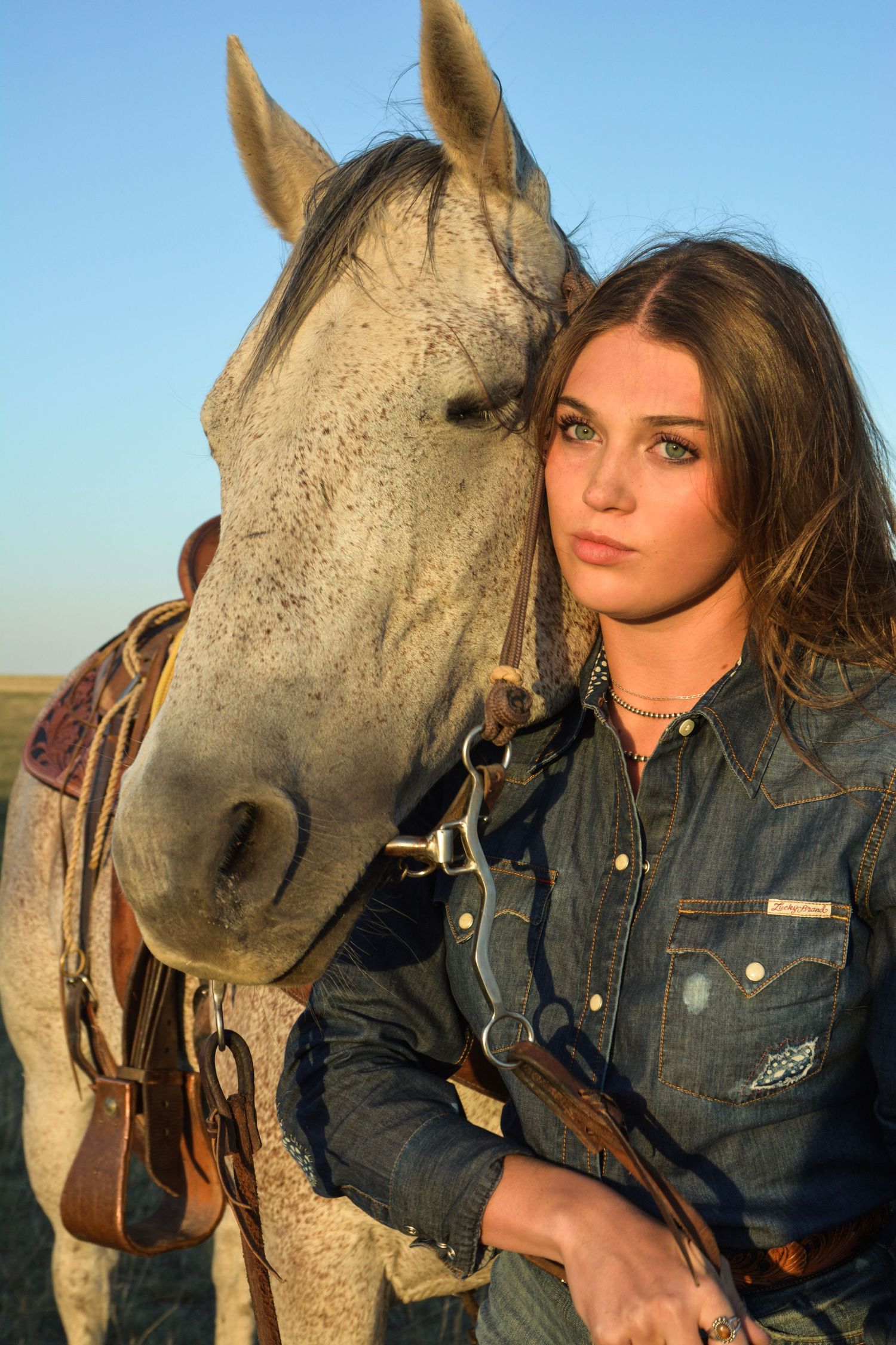 Young woman in denim shirt poses with a gray horse outdoors during golden hour on open plains.