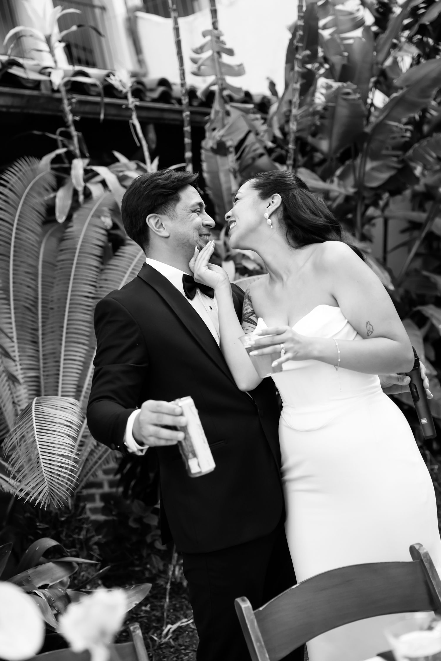 Black and white photo of a laughing bride in strapless gown touching groom's face outdoors amid tropical plants.