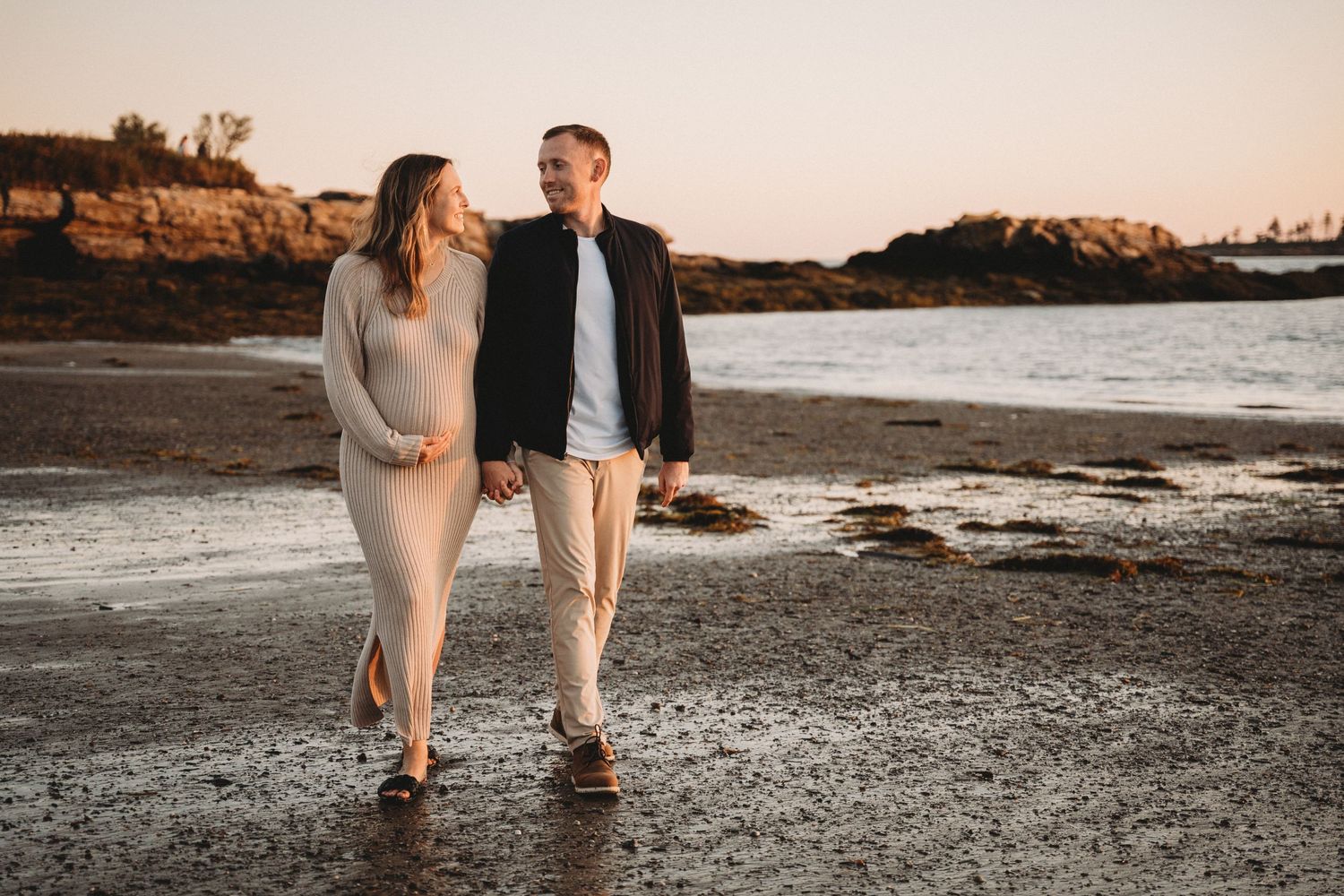 Pregnant couple walking hand-in-hand on a rocky beach at sunset, wearing casual outfits with golden light behind them.