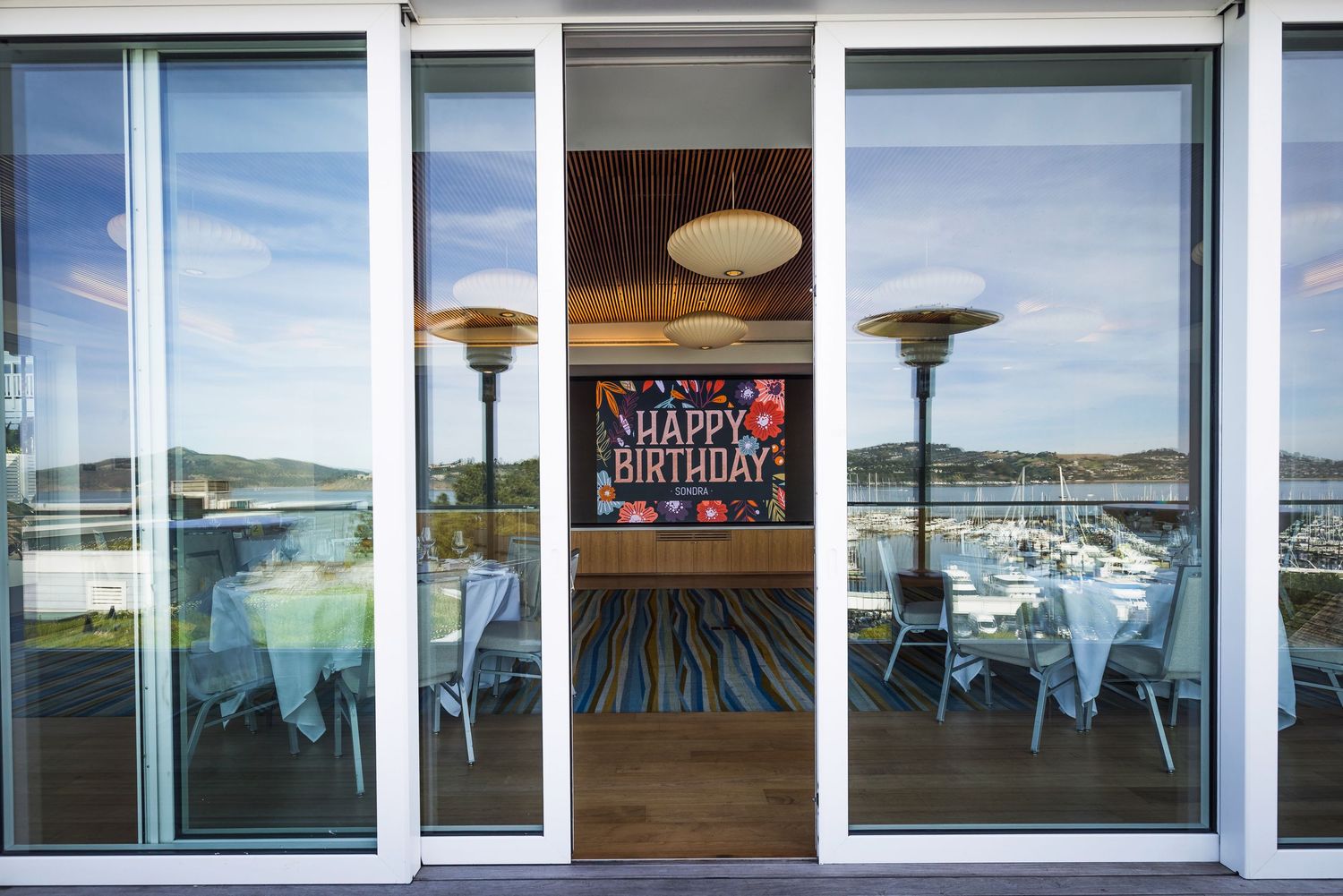 Indoor dining area with Happy Birthday banner, patio heaters, and marina views through large glass sliding doors.