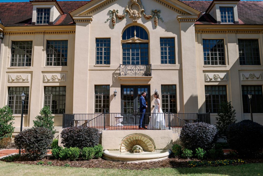 Bride and groom pose in front of an elegant beige mansion with a fountain inside Turtle Court at the Thomas Center in Gainesville, Florida.