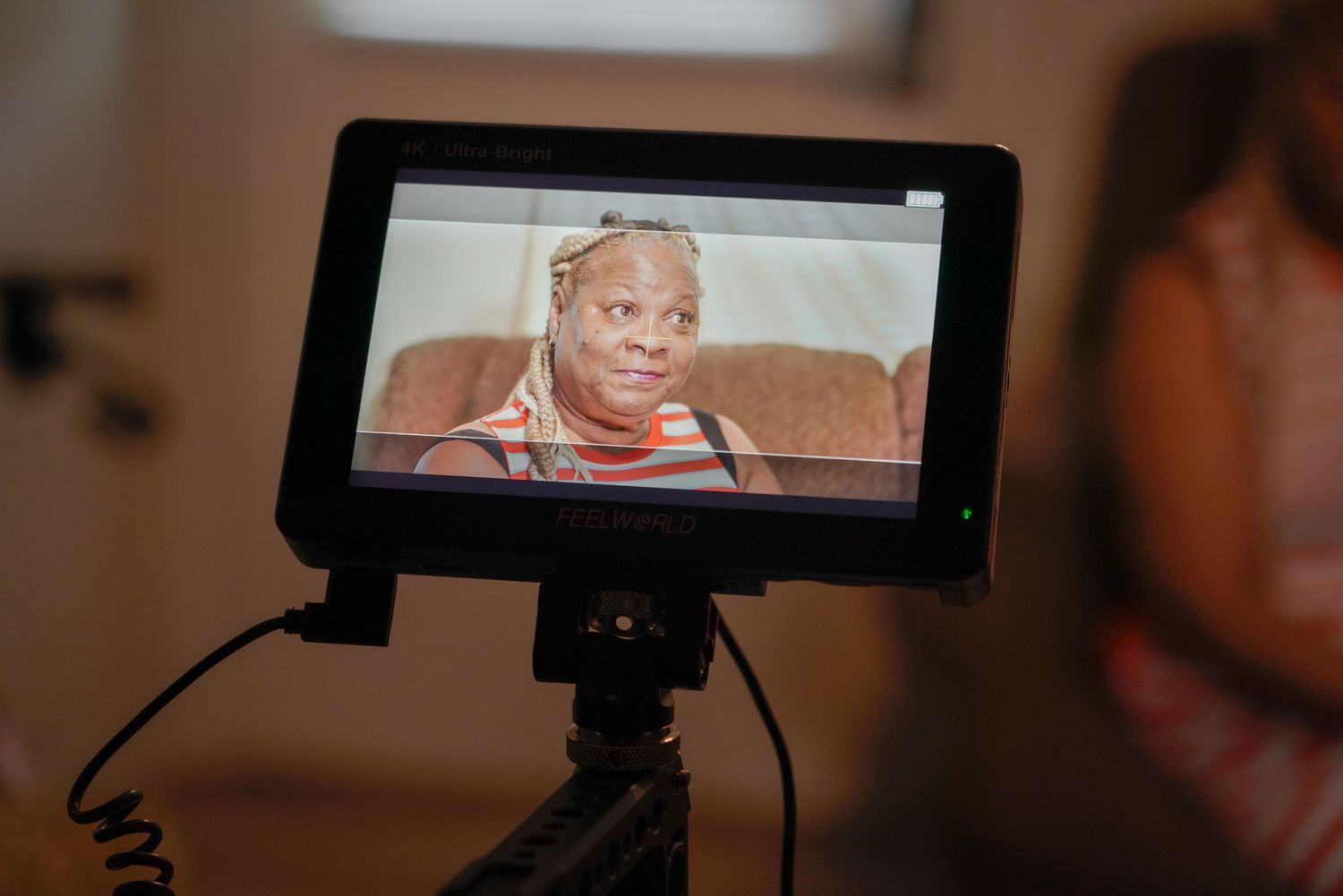 Camera monitor displaying an elderly woman being filmed during a video interview in a warm indoor setting.