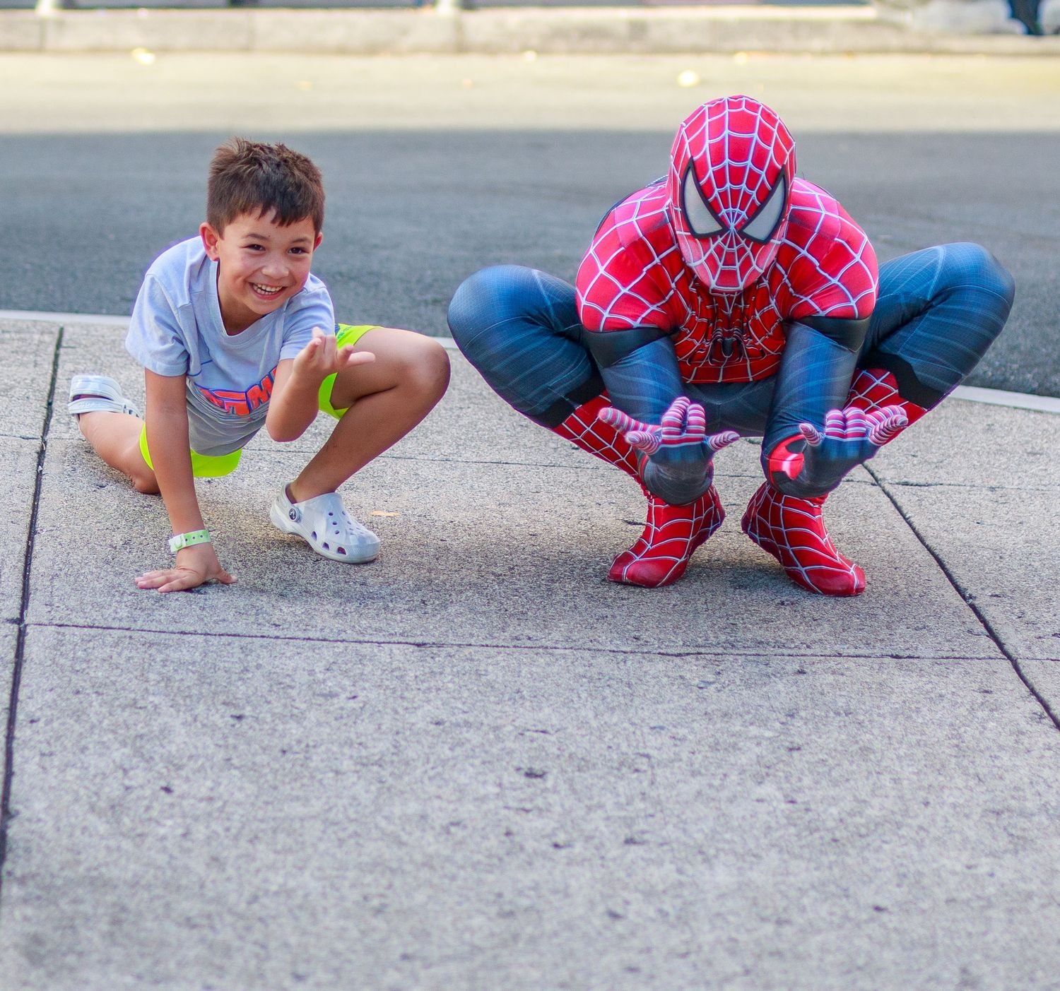 A young boy and person in Spiderman costume crouch together on a sidewalk, smiling for a photo.