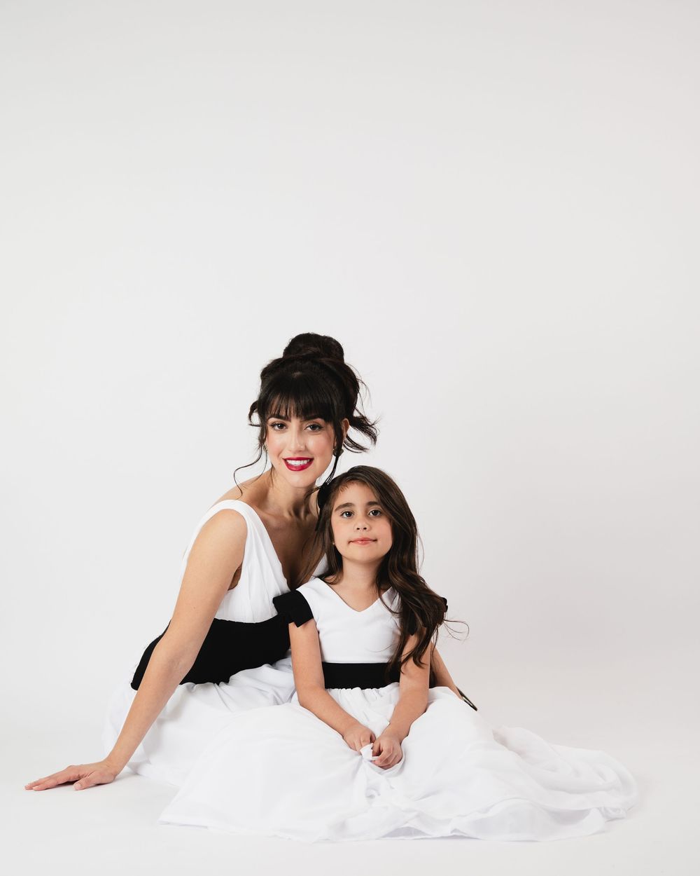 A woman and young girl in matching white and black dresses pose together on a white studio background.