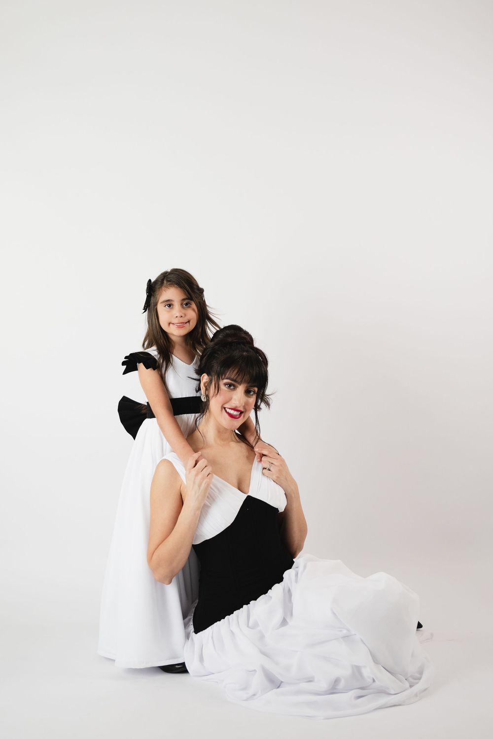 A woman in a black and white gown sits on the floor while a young girl in a matching dress stands behind her.