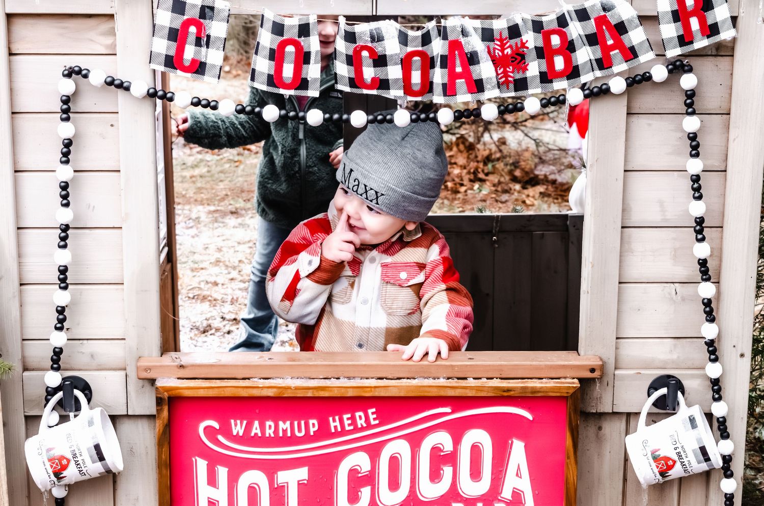 Toddler in plaid shirt at a festive hot cocoa bar stand decorated with holiday garland.