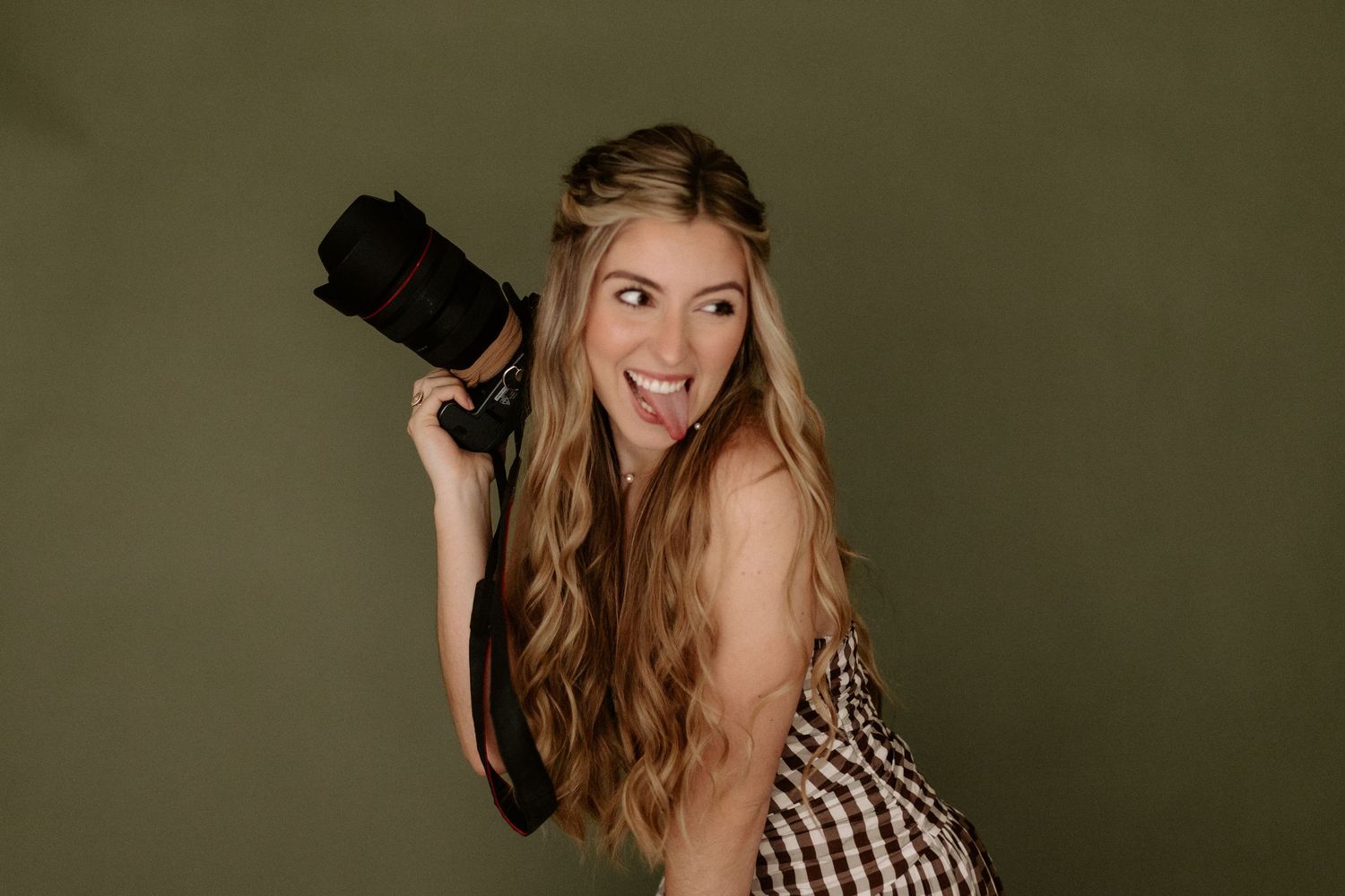 Young woman with long blonde hair holding a DSLR camera, smiling playfully against an olive green background.