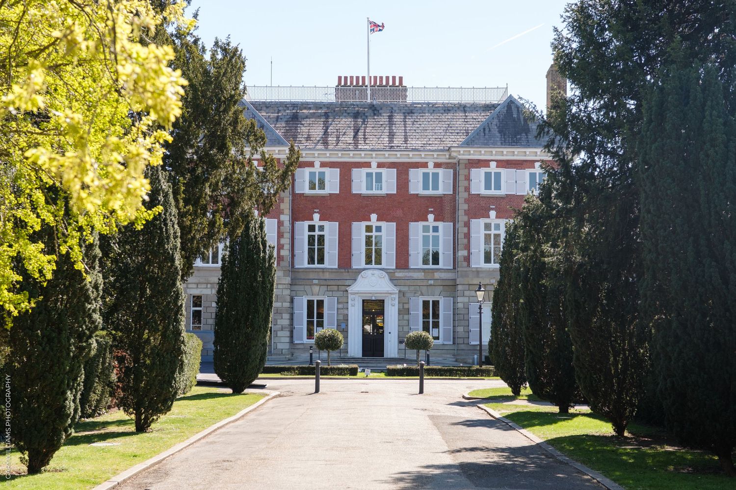 Grand red-brick Georgian manor house with symmetrical facade, flanked by tall cypress trees lining a formal driveway.