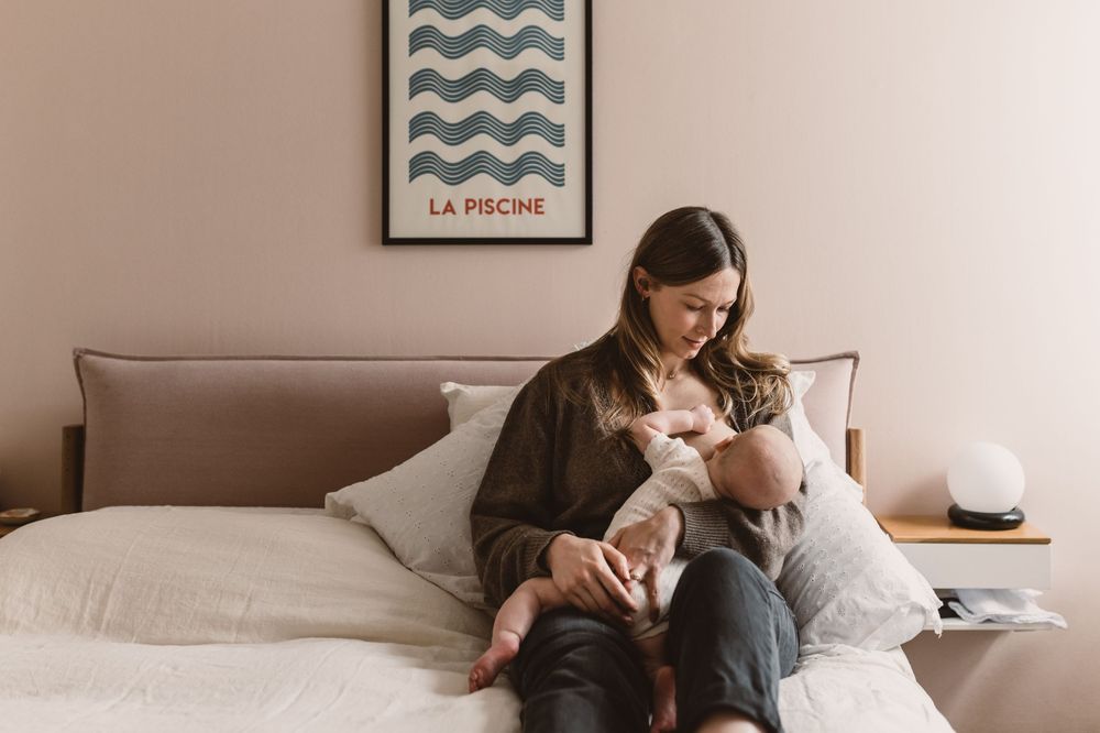 Mother breastfeeding newborn baby on bed with pink walls and art print in cozy bedroom.