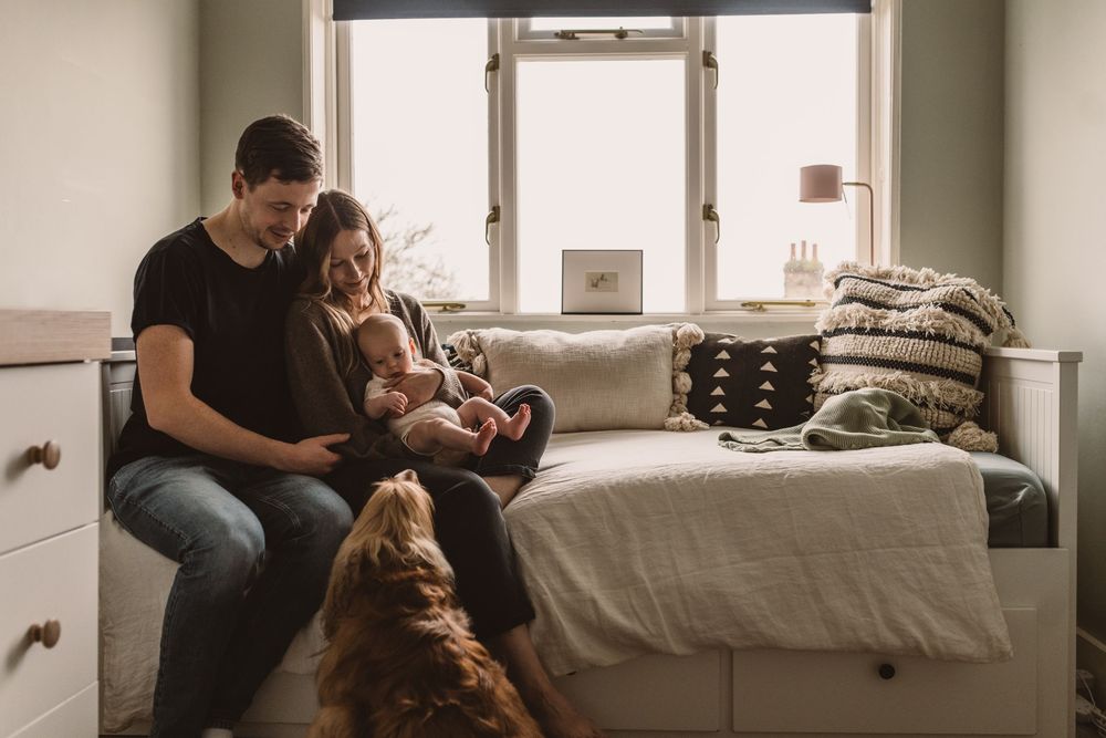 Young couple sitting on a daybed with their newborn baby while their dog looks up.