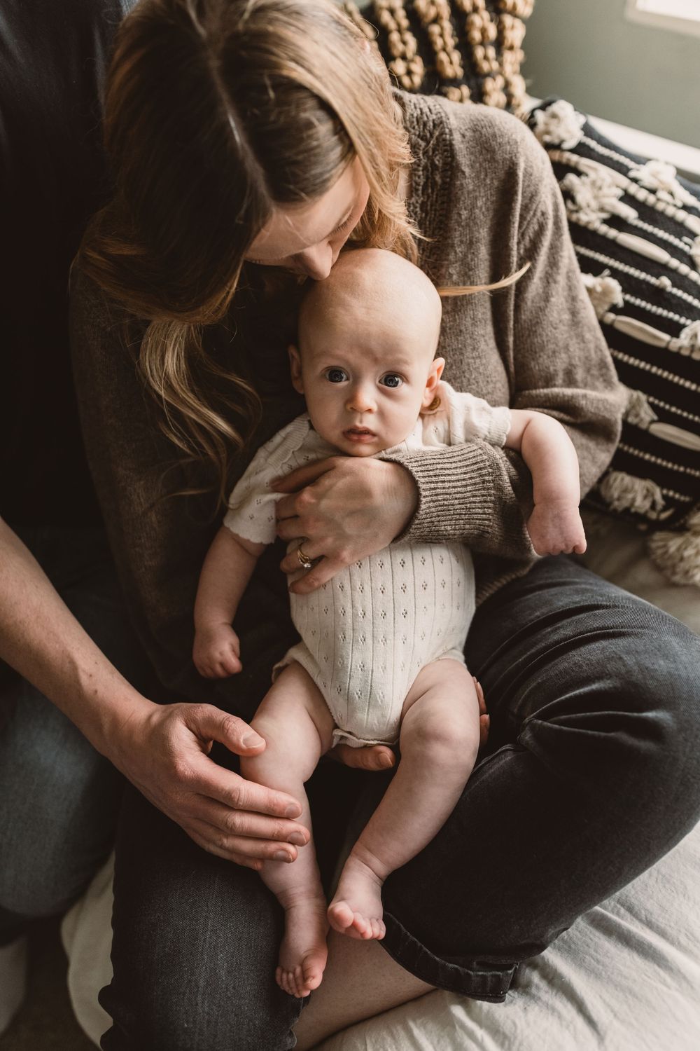 A mother tenderly holds her alert baby in her lap while seated on a bed with cozy textured pillows.