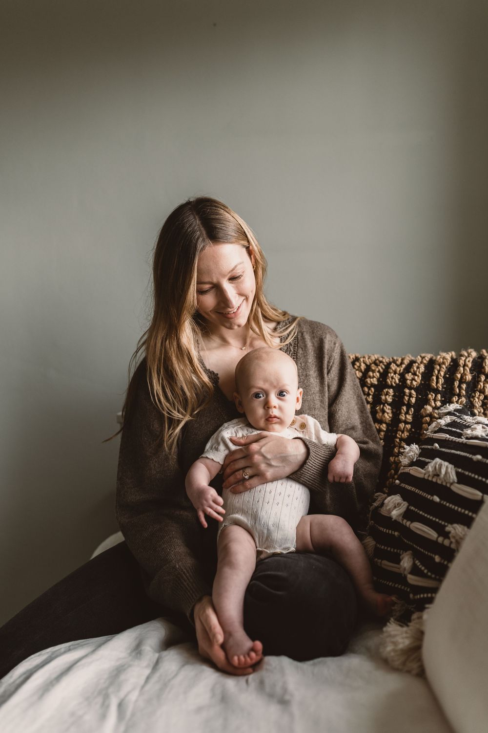 Smiling mother with long hair holding her baby in a cozy knit sweater, sitting on a white bed.
