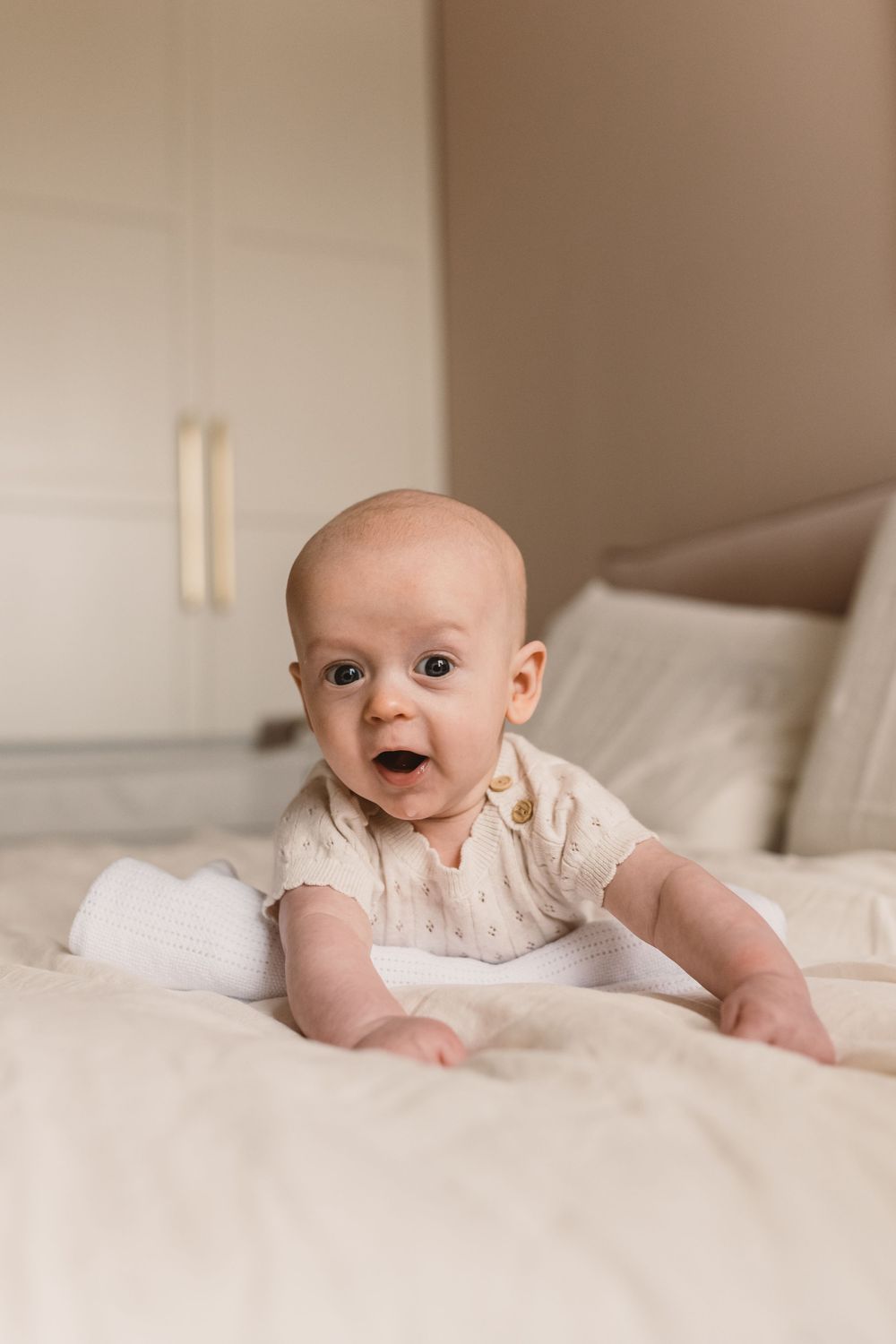 13-week-old baby in cream onesie doing tummy time on white bed, looking up with wide eyes and open mouth.