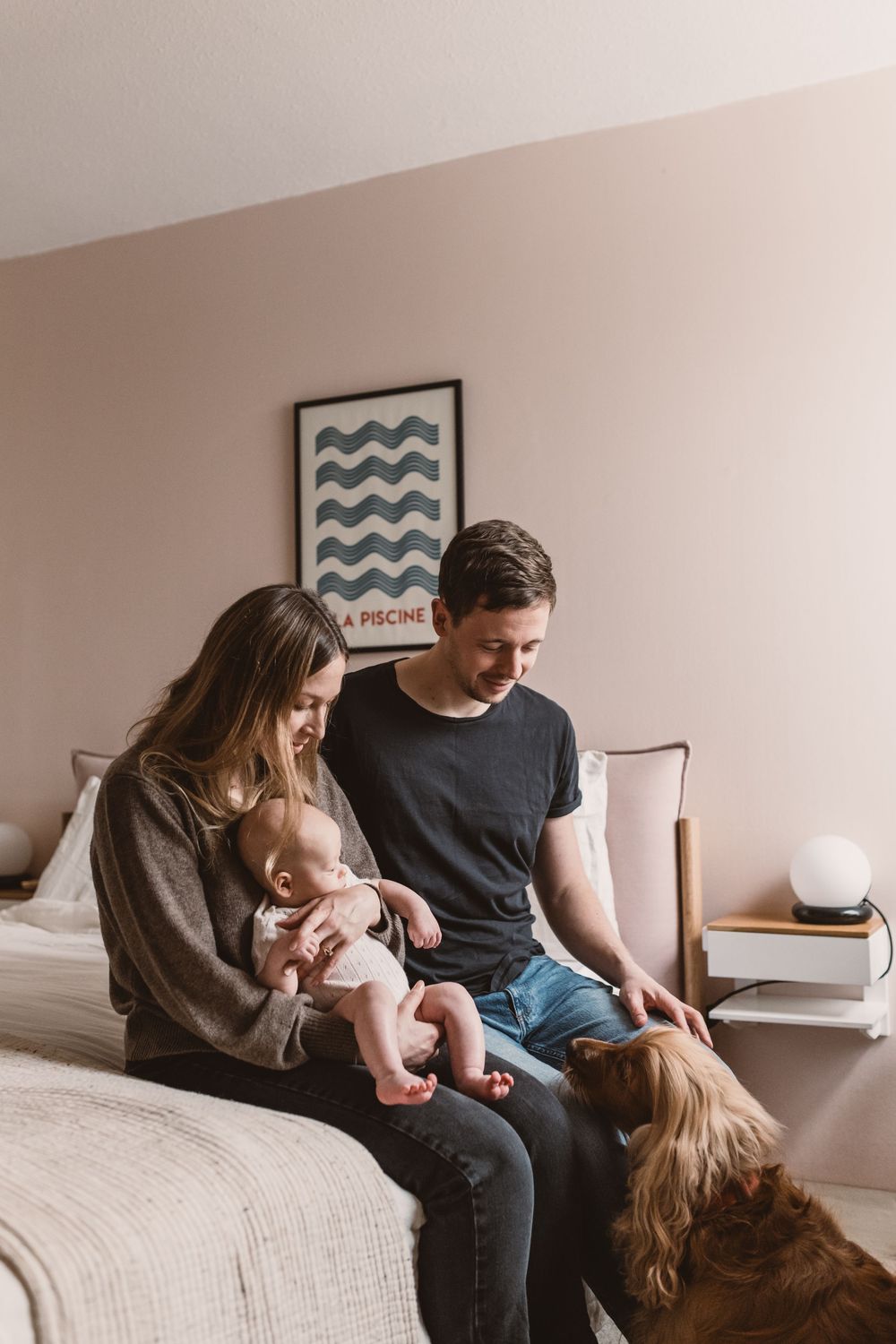 Young couple sitting on bed with baby and dog in cozy pink bedroom with an art print.