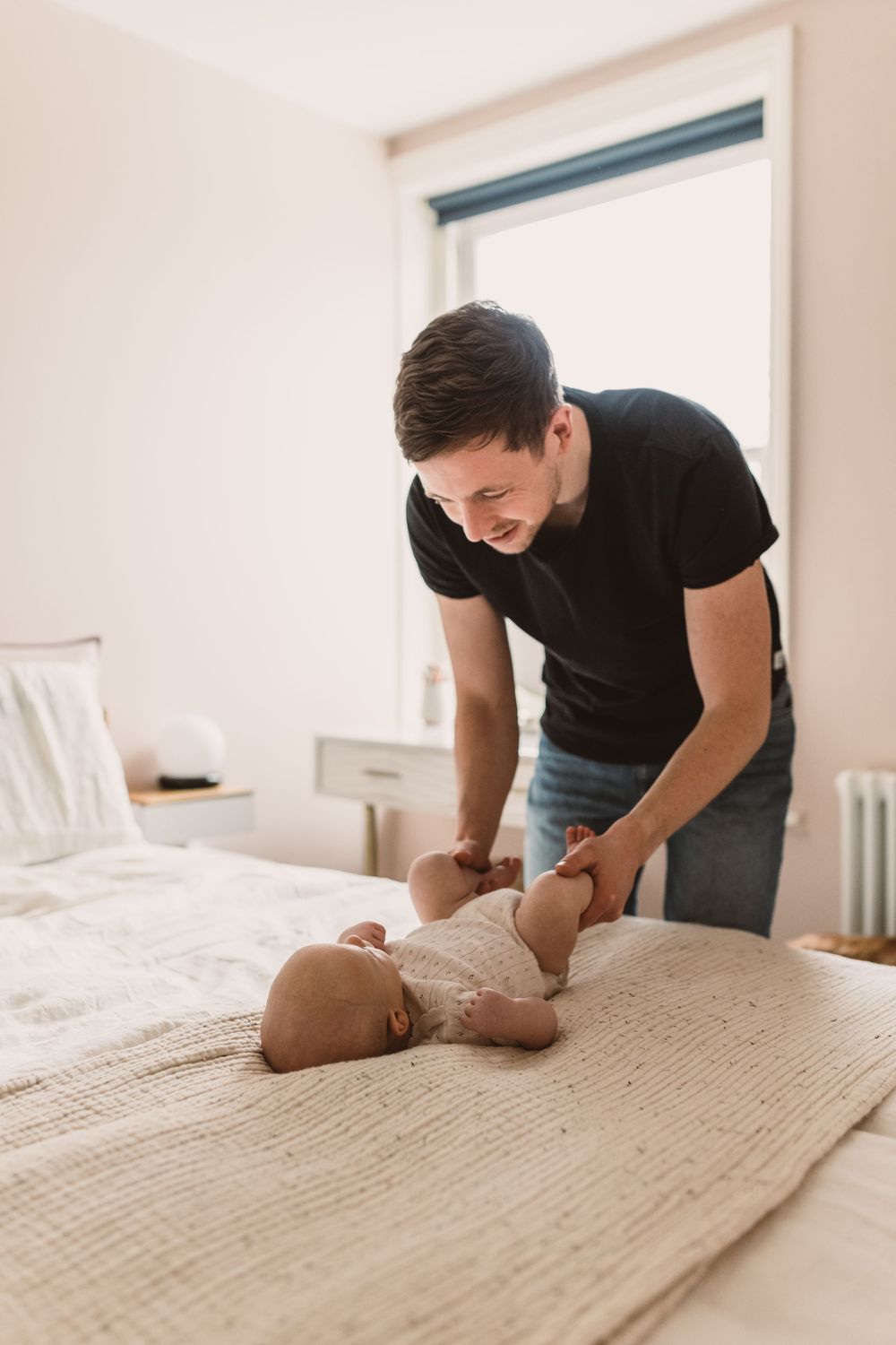 Father in black t-shirt carefully tending to newborn baby lying on bed in bright, minimalist bedroom.