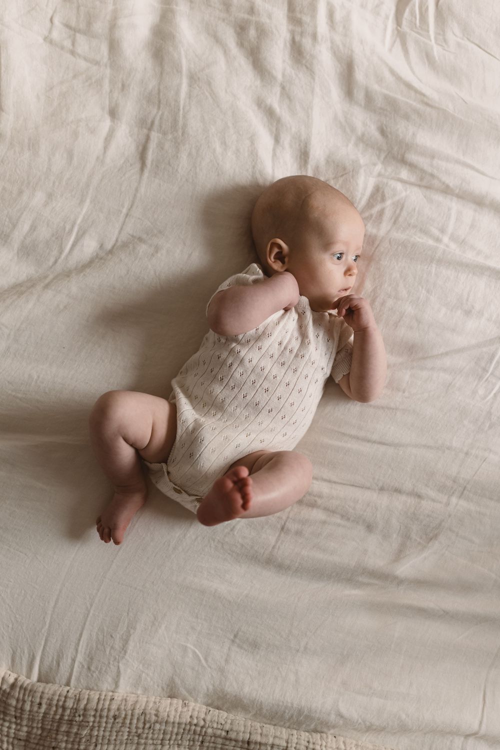 A peaceful baby in a cream knit onesie lies on a soft white linen blanket, gazing to the side.