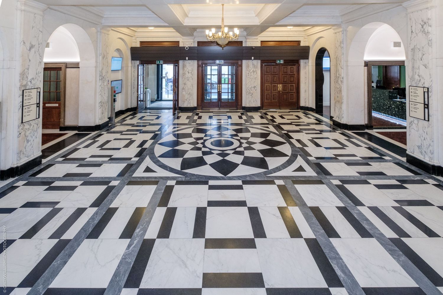 Elegant marble lobby featuring a stunning black and white geometric floor mosaic, arched doorways, and a grand chandelier.