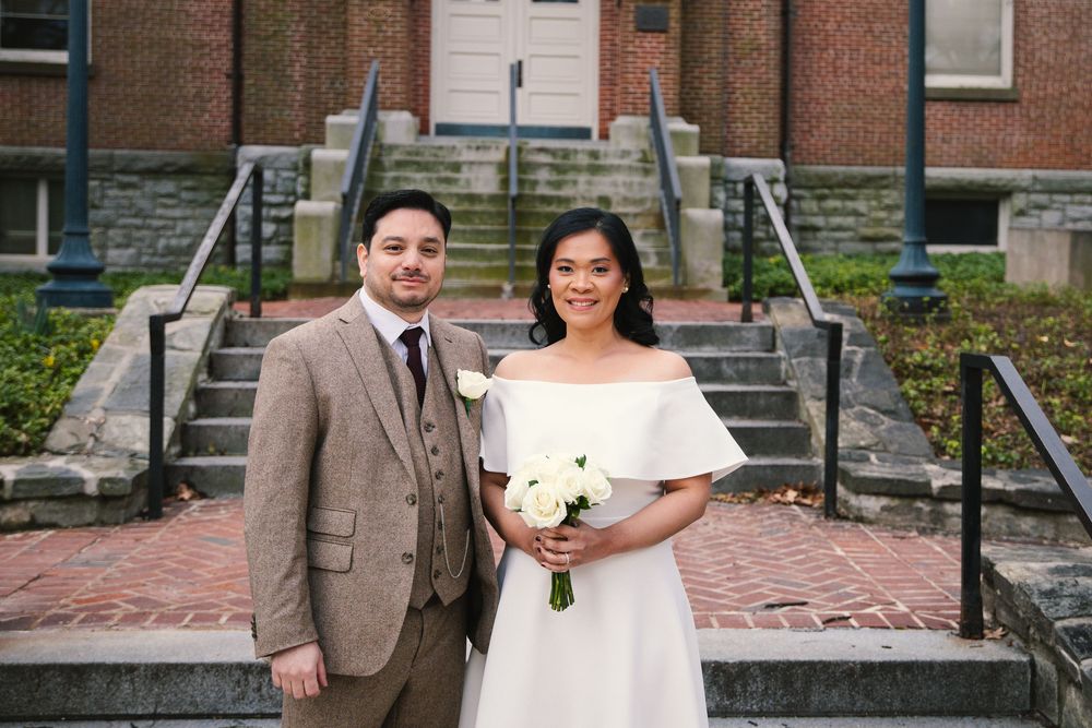 Bride and groom posing together in front of brick building steps, bride holding white bouquet in elegant off-shoulder wedding dress.