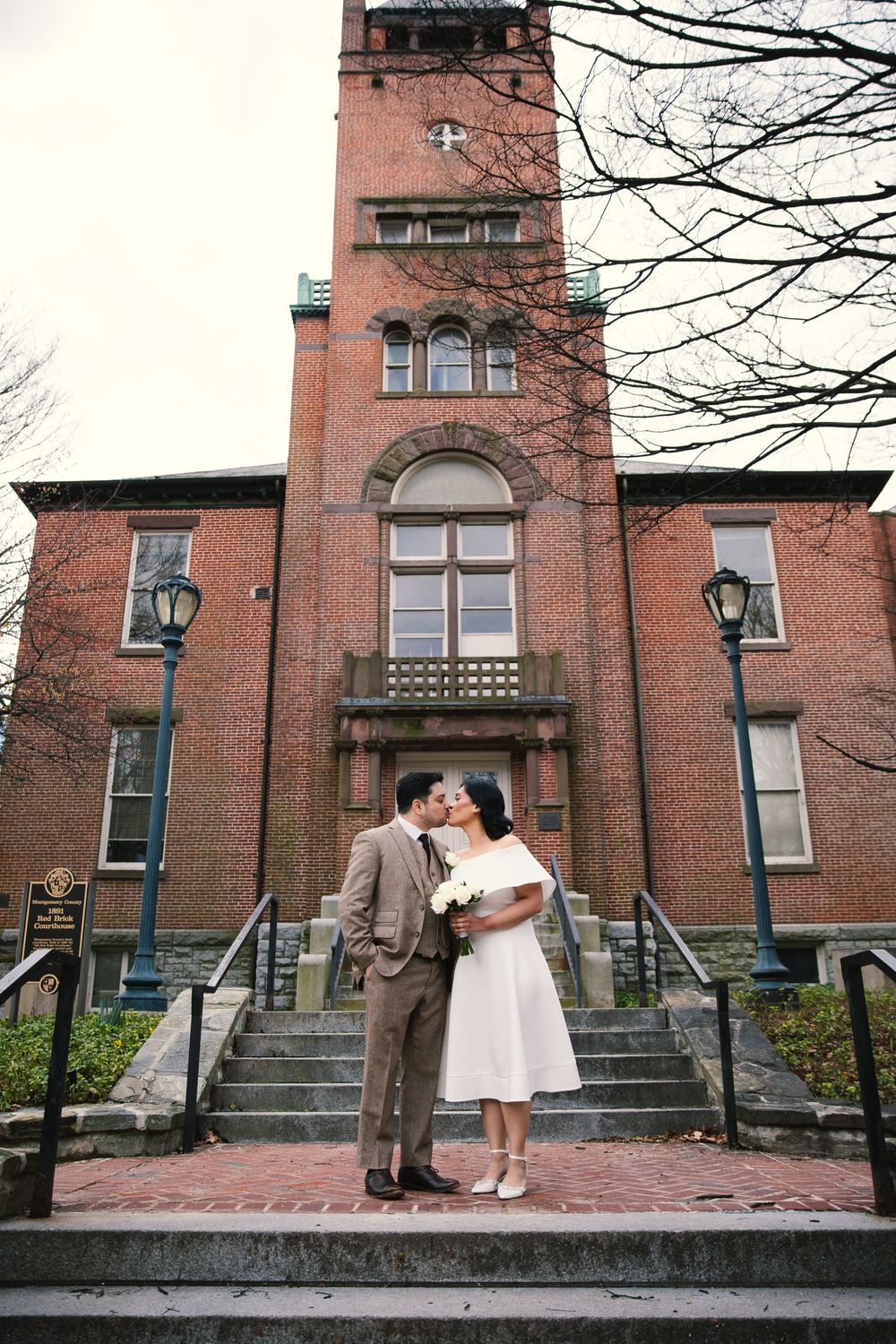 Bride and groom kissing on steps of red brick church, she holds white bouquet, he wears brown tweed suit.