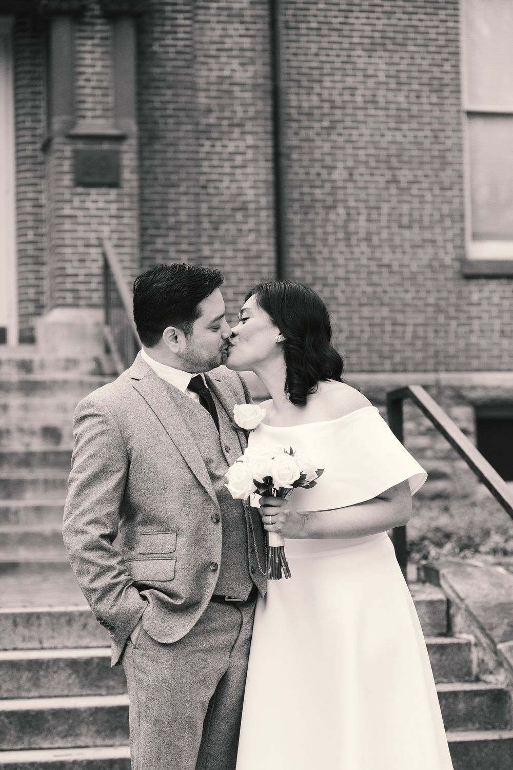 Black and white photo of a bride and groom sharing a kiss outside a brick building, bride holding white bouquet.