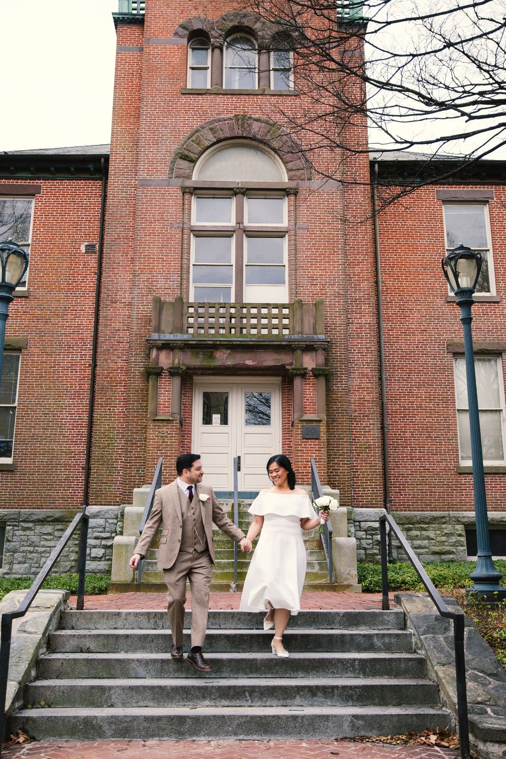 Bride and groom walk down steps of historic brick building, she in white dress, he in tan suit, holding hands.