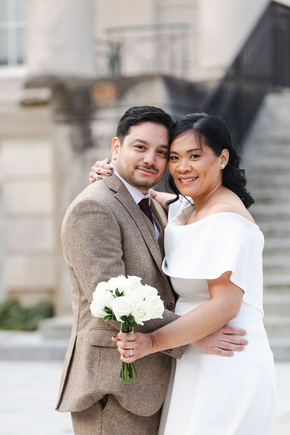 Happy interracial couple embracing outdoors on wedding day, bride in white off-shoulder dress holding white roses.