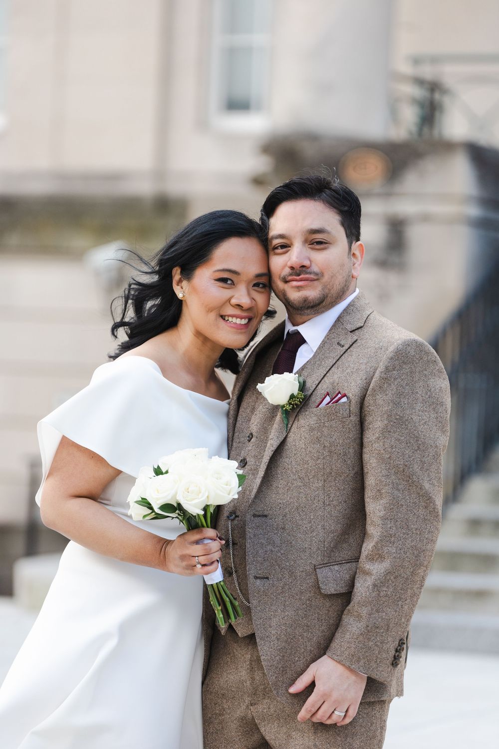 Smiling bride in white dress holding white roses bouquet poses with groom in brown tweed suit outside elegant building.