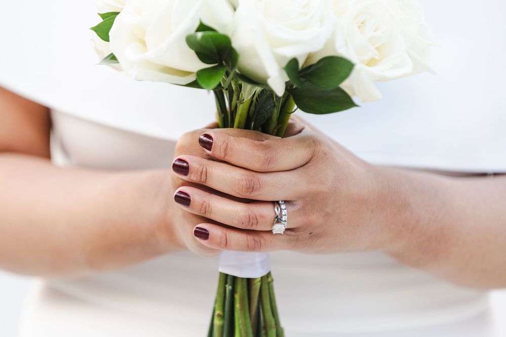 Bride holding white rose bouquet with diamond engagement ring and dark burgundy nail polish on wedding day.