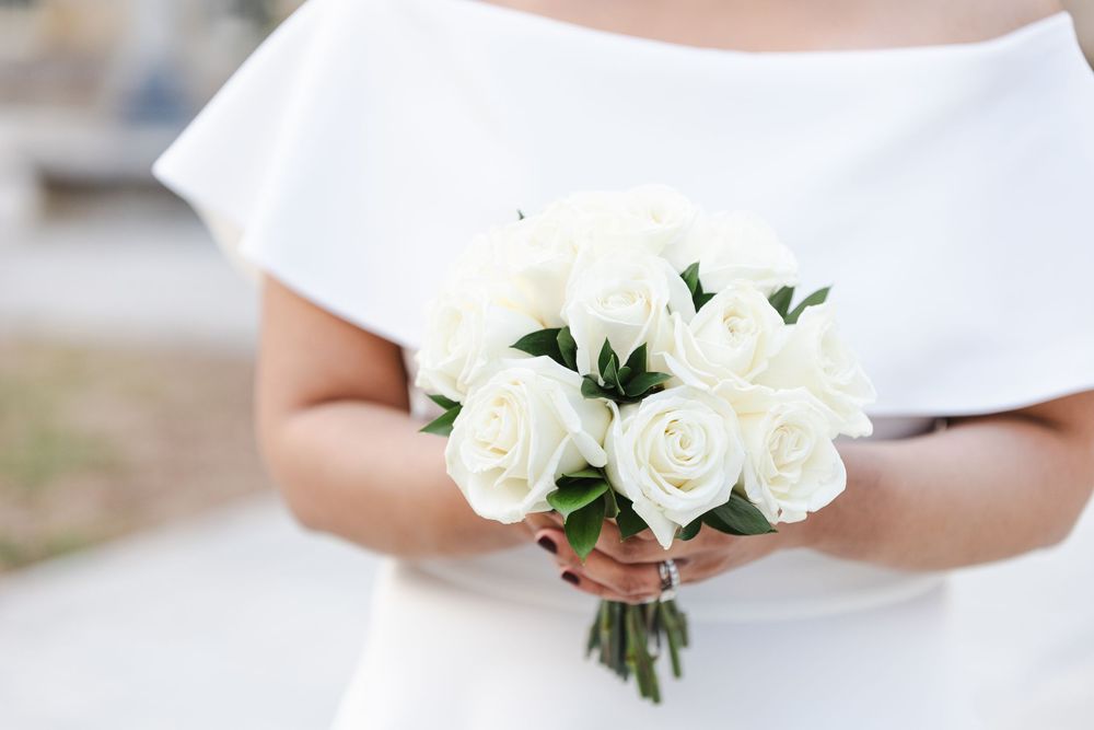 Bride in white off-shoulder dress holding elegant bouquet of white roses at wedding ceremony.