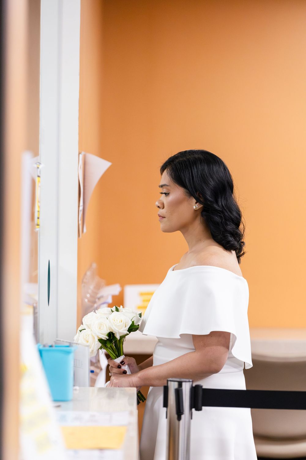 Elegant bride in white off-shoulder dress holding white flower bouquet standing at a counter with orange wall background.