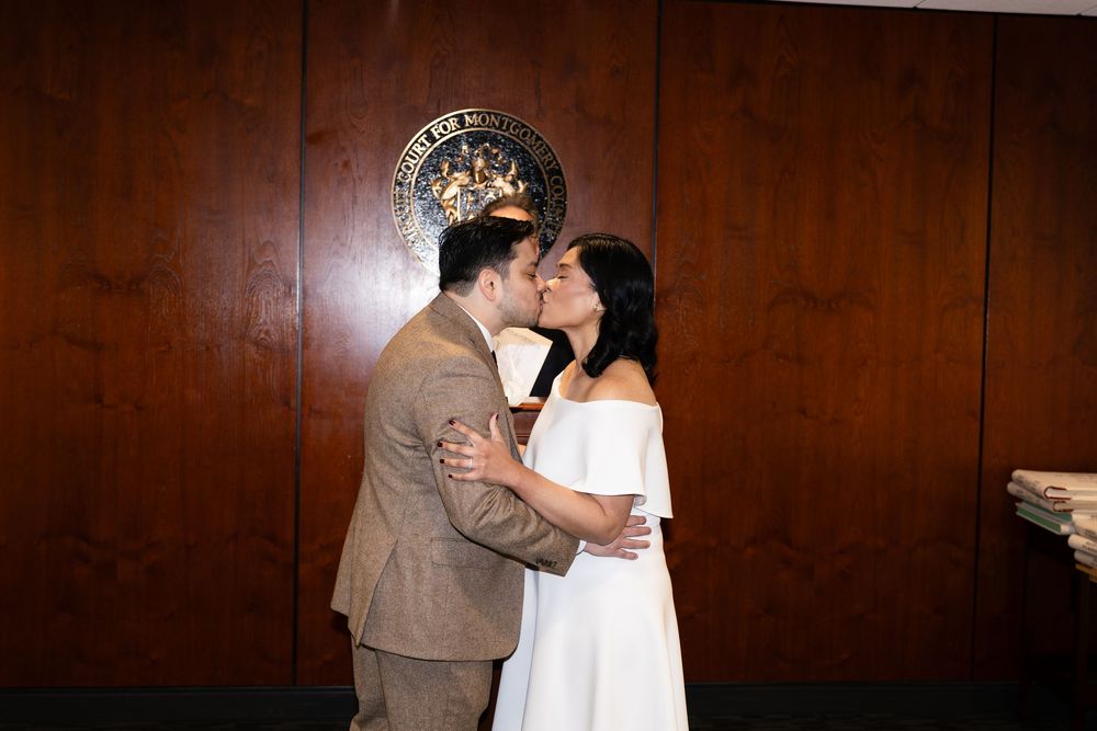 Couple sharing first kiss at courthouse wedding ceremony, groom in tan suit, bride in white off-shoulder dress.