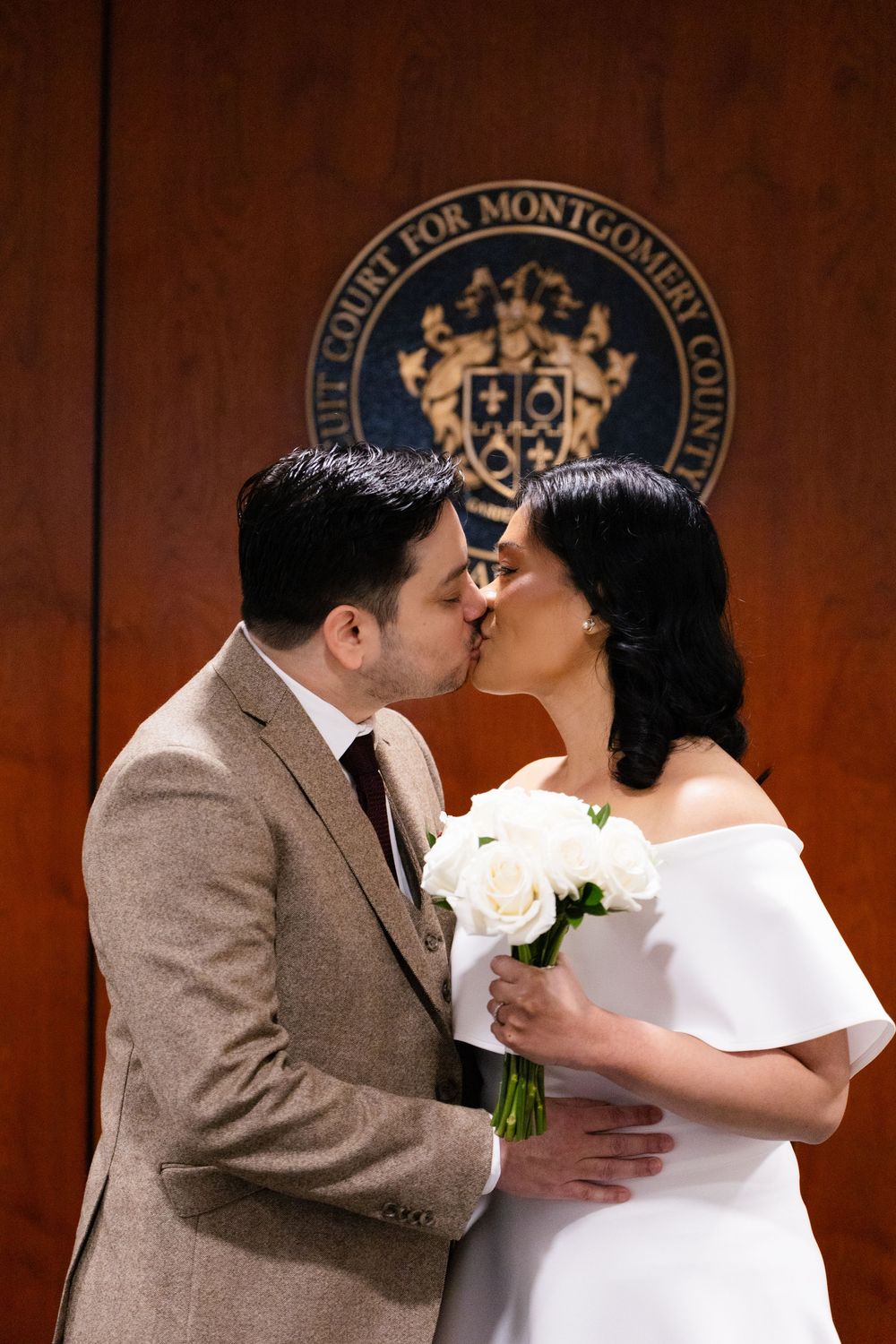 Couple sharing first kiss at Montgomery County Circuit Court wedding ceremony, bride in white dress holding white rose bouquet.
