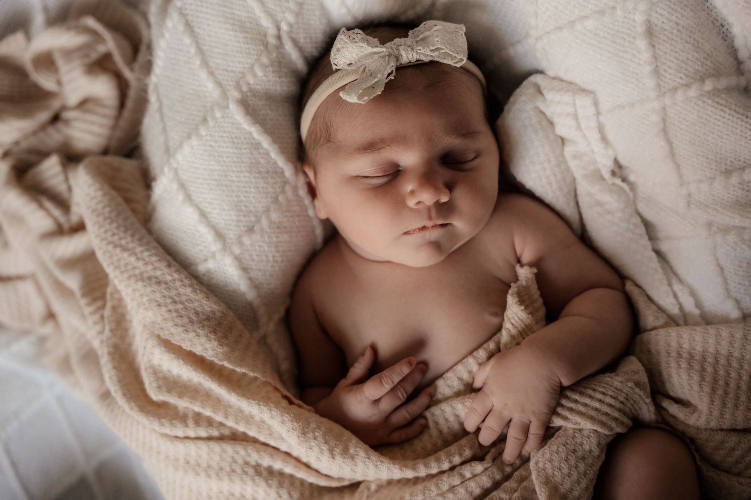 Peaceful newborn baby girl sleeping wrapped in a soft beige knit blanket with a bow headband.