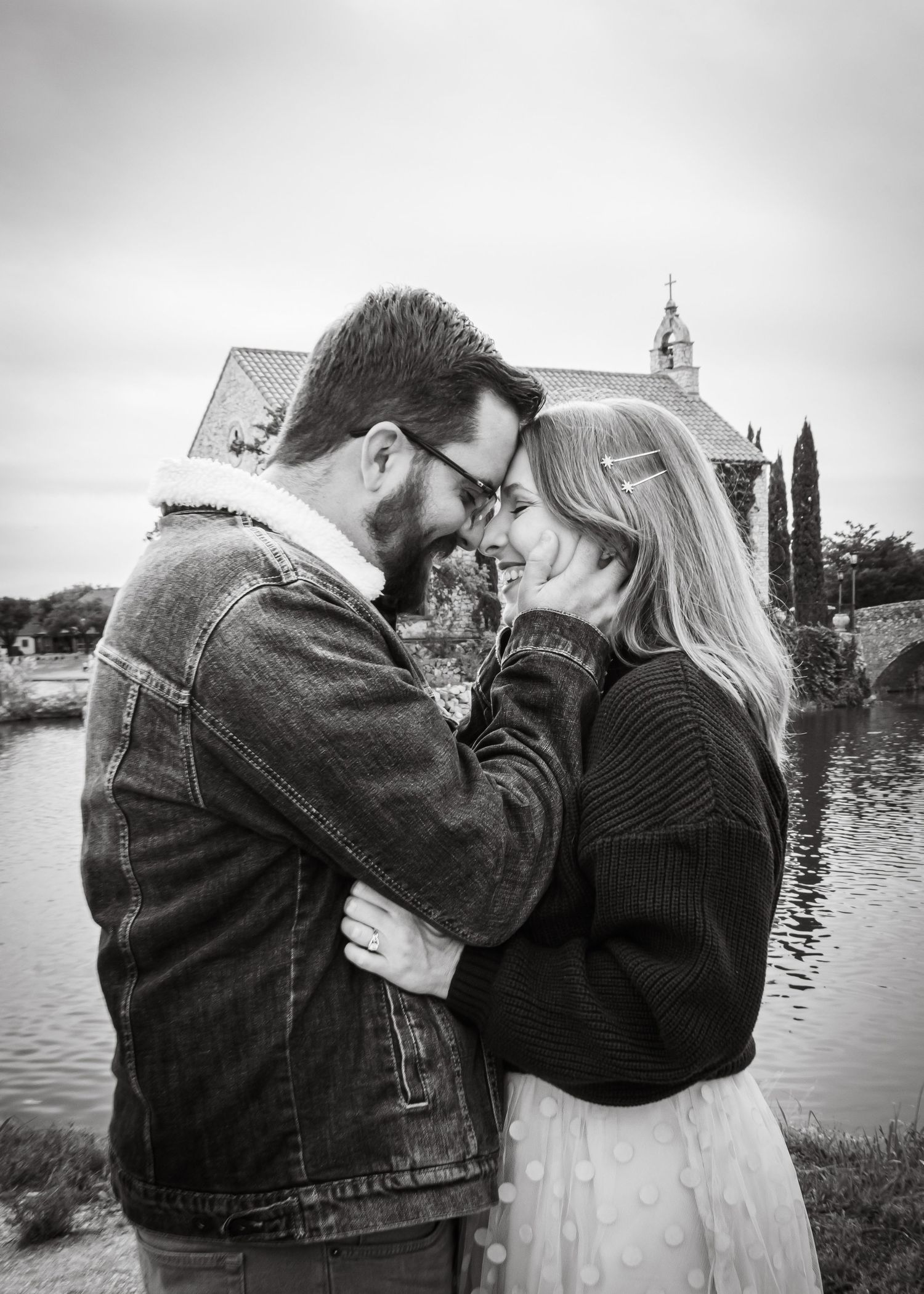 A black and white photo of a couple embracing and smiling near a scenic waterfront.