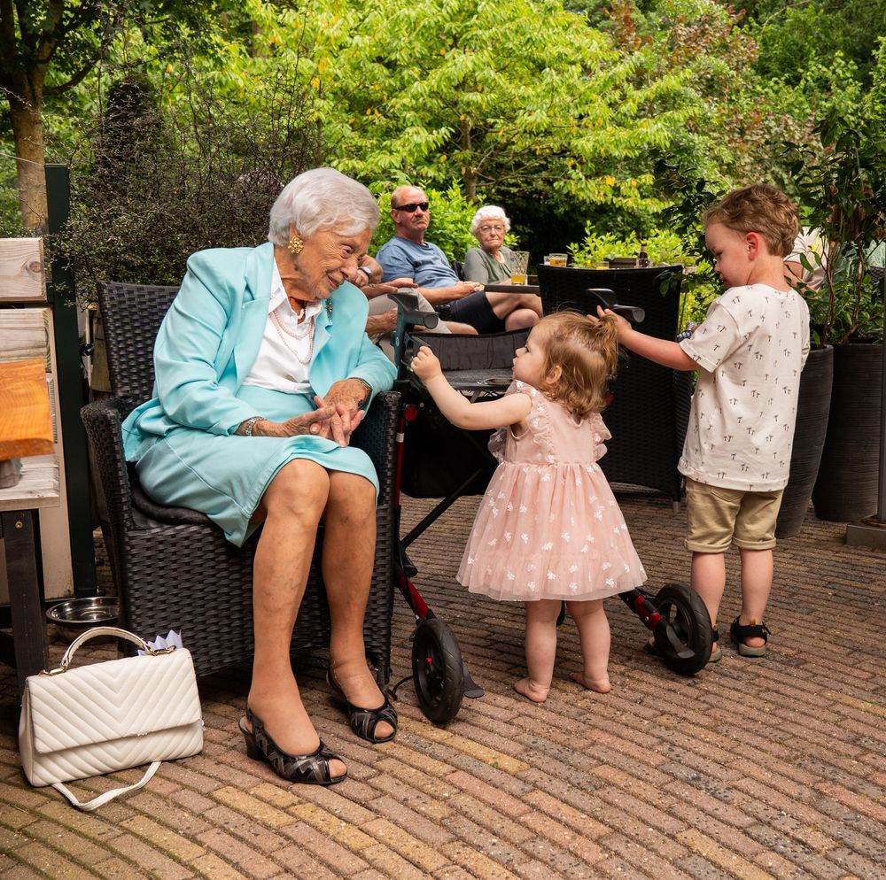 Een oudere vrouw in een turquoise outfit speelt met twee jonge kinderen op een terras. documentaire fotograaf