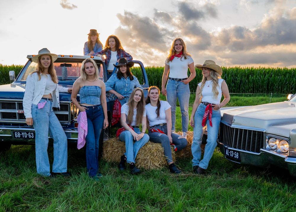 Een groep van negen vrouwen in westerse kleding poseert bij oldtimers en hooibalen tijdens zonsondergang.