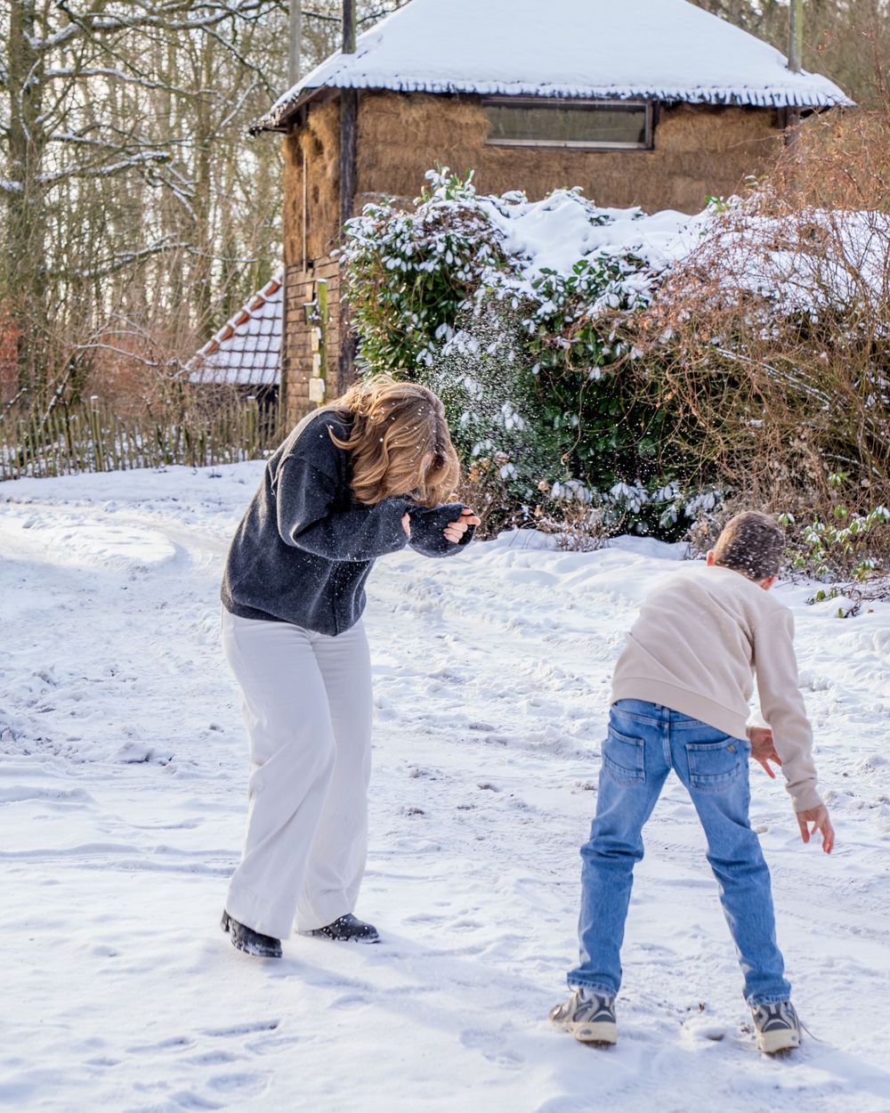 Moeder en kind spelen in de sneeuw buiten een rustiek huis, omgeven door winterse bomen.