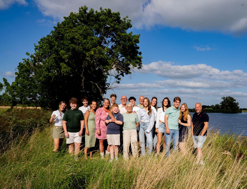 Groot familieportret, staand in hoog gras bij een meer onder een blauwe hemel.