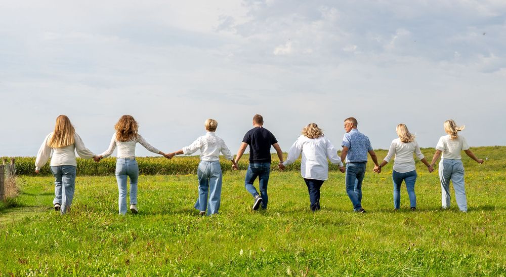 Een groep van acht mensen loopt hand in hand door een groene weide onder een bewolkte hemel. familie fotograaf