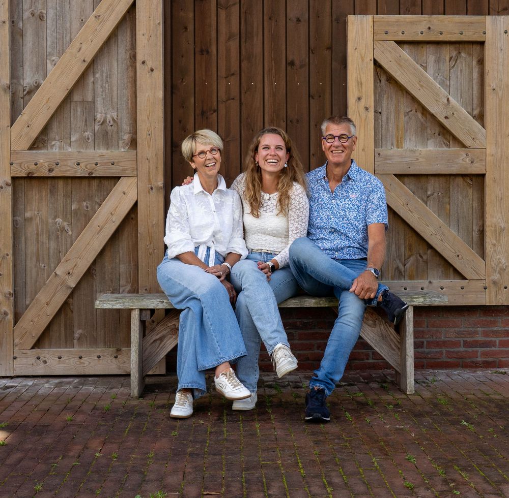 Een lachend gezin van drie zit op een houten bankje voor grote schuurdeuren buiten.